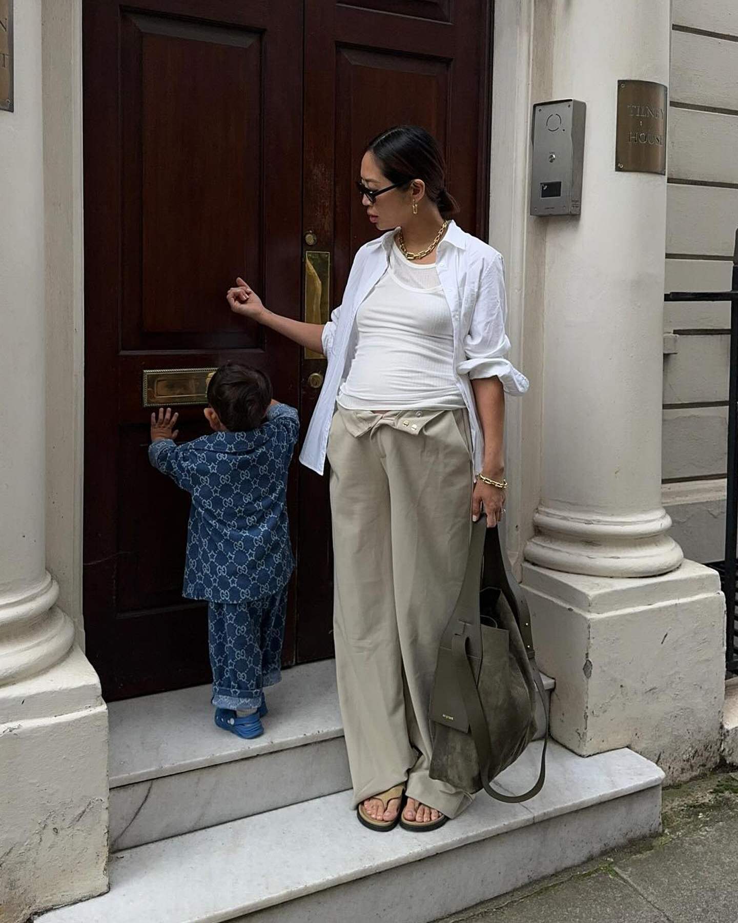 A pregnant Aimee Song poses with her son wearing a chic maternity outfit with layered white tank tops, a white button-down shirt, folded waistband tan pant, a green designer bag, and tan suede flip-flop sandals