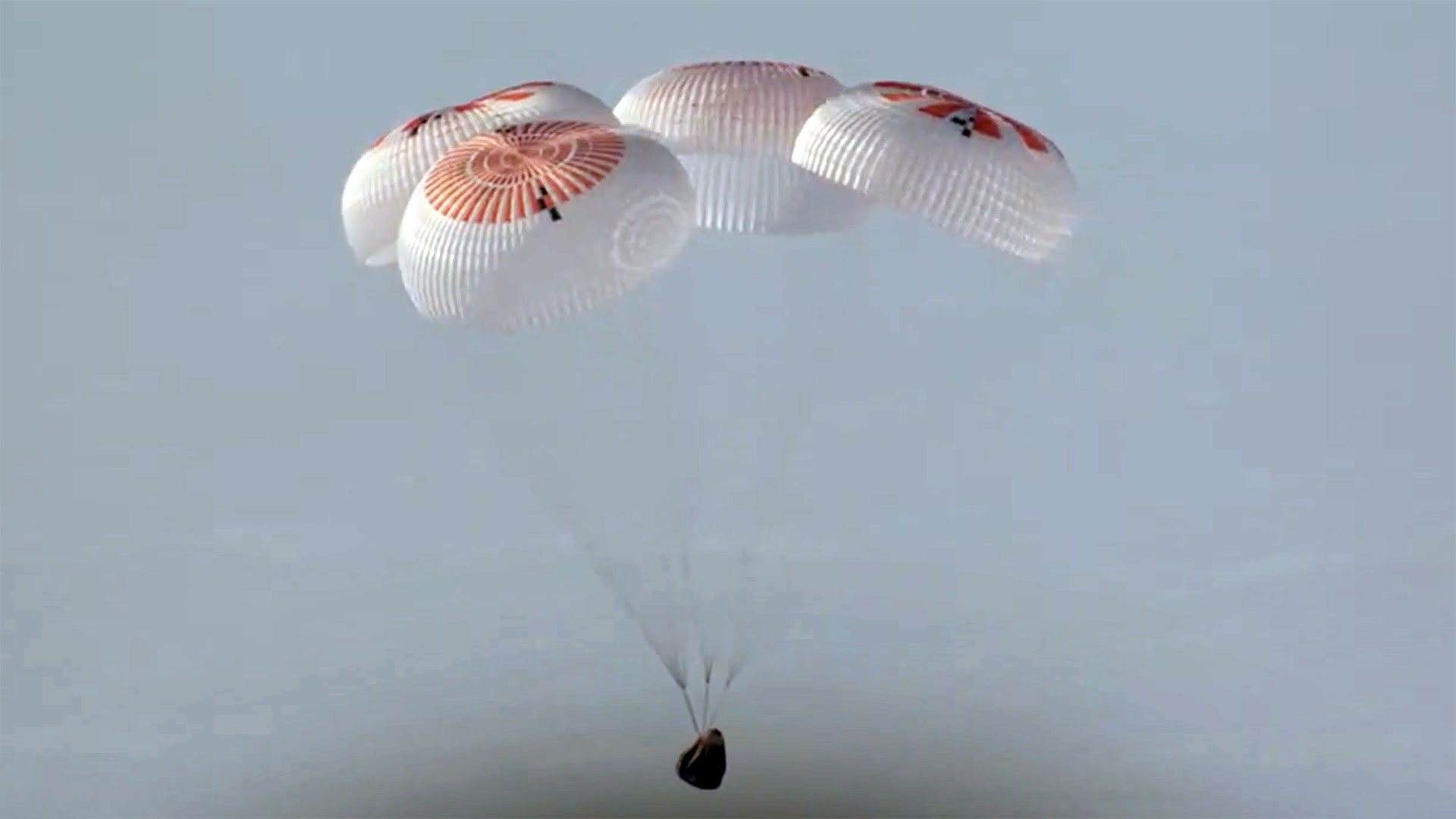 A capsule descends toward an ocean splashdown under four orange and white parachutes