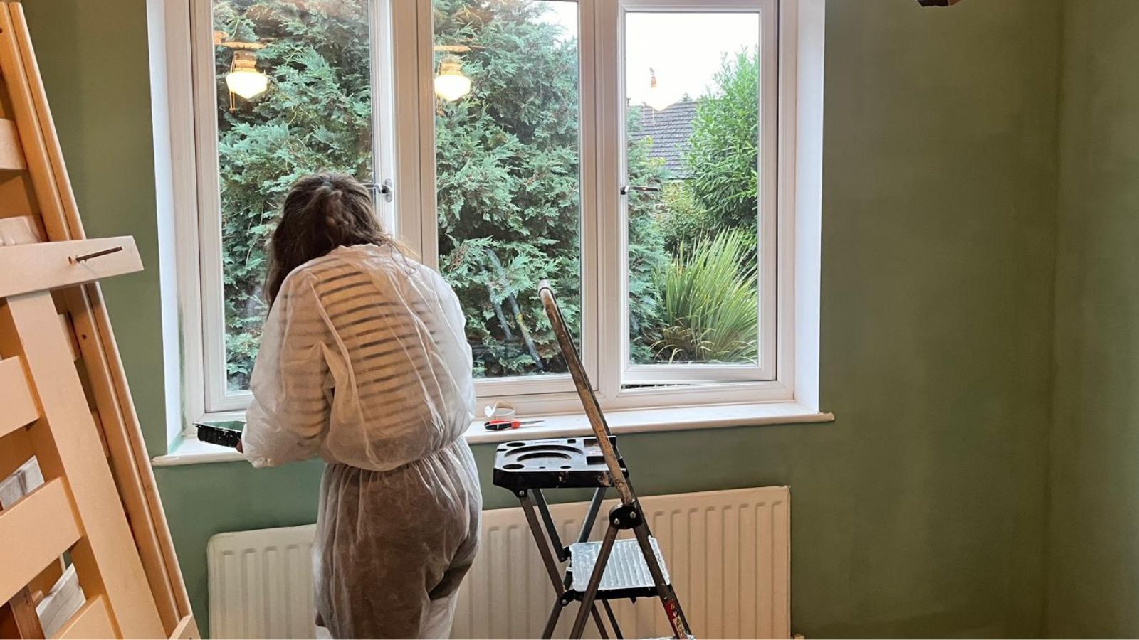 A person in a white disposable decorating suit paints window frames in a room with freshly painted sage green walls, a stepladder and paint tray beside them.