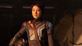 Still from the Star Wars T.V. Show Star Wars The Mandalorian. Here we see Sabine Wren with dark purple short hair and brown eyes. She is wearing special Mandalorian armor and is holding her helmet in the crook of her arm.