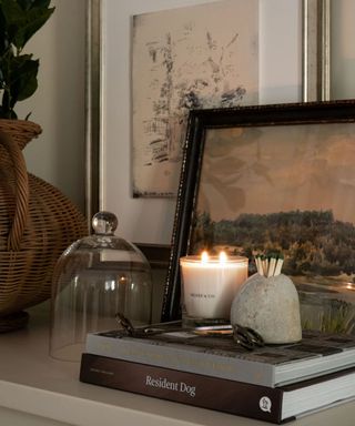 entryway console vignette styled with vintage artwork a rattan vase and a stack of books with a scented candle and matches holder