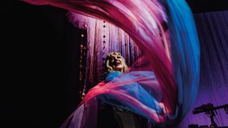 A performer twirls vibrant pink and blue fabrics on stage, surrounded by shimmering backdrop decorations and colorful lighting