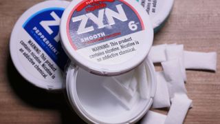 Three circular containers of Zyn nicotine pouches are shown on a wooden table. The container on top is open and nicotine pouches are spilling out onto the table. 