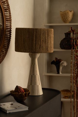 A lamp with a handwoven rattan shade sits on top of a black cabinet before a shelf unit filled with similarly inclined objects.