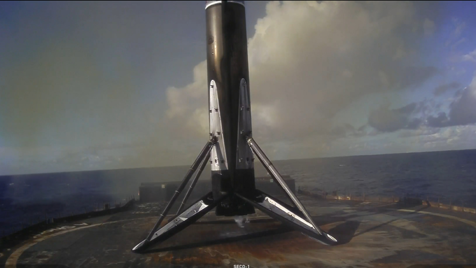 a charred rocket stage stands on legs on a barge at sea