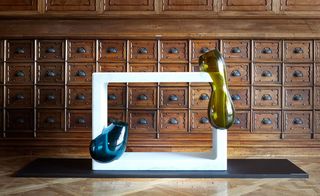 A marble hollow square with two glass shapes built into it in front of a wooden chest of drawers..