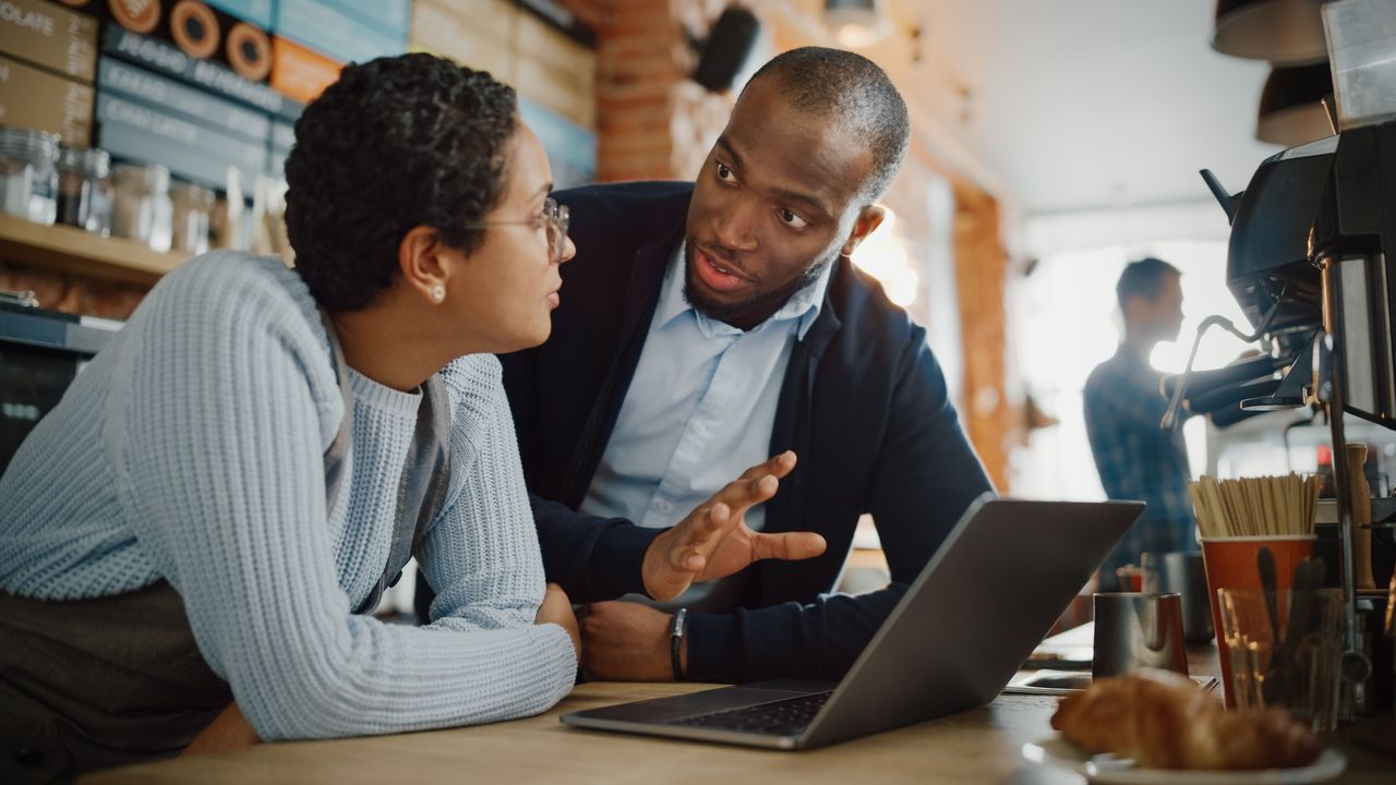 A small business owner talks seriously with an employee while they&#039;re looking at a laptop together in a coffee shop.