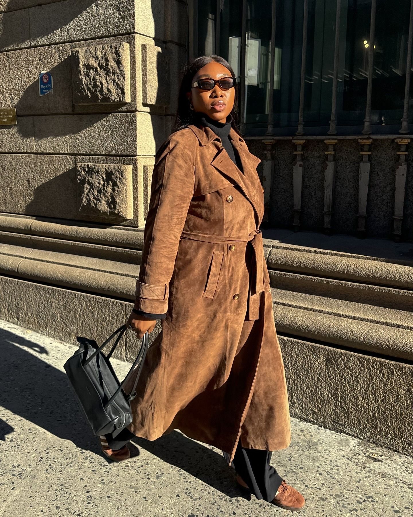 Influencer Nnenna wears a suede long coat, black East West bag, black slacks, and suede Adidas sneakers.