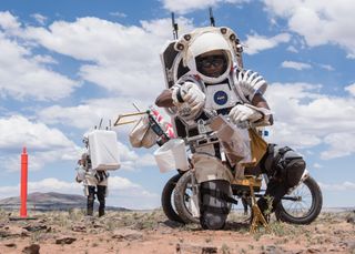 two people in skeleton-like spacesuits without the outer shell walk through the desert under a bright sun