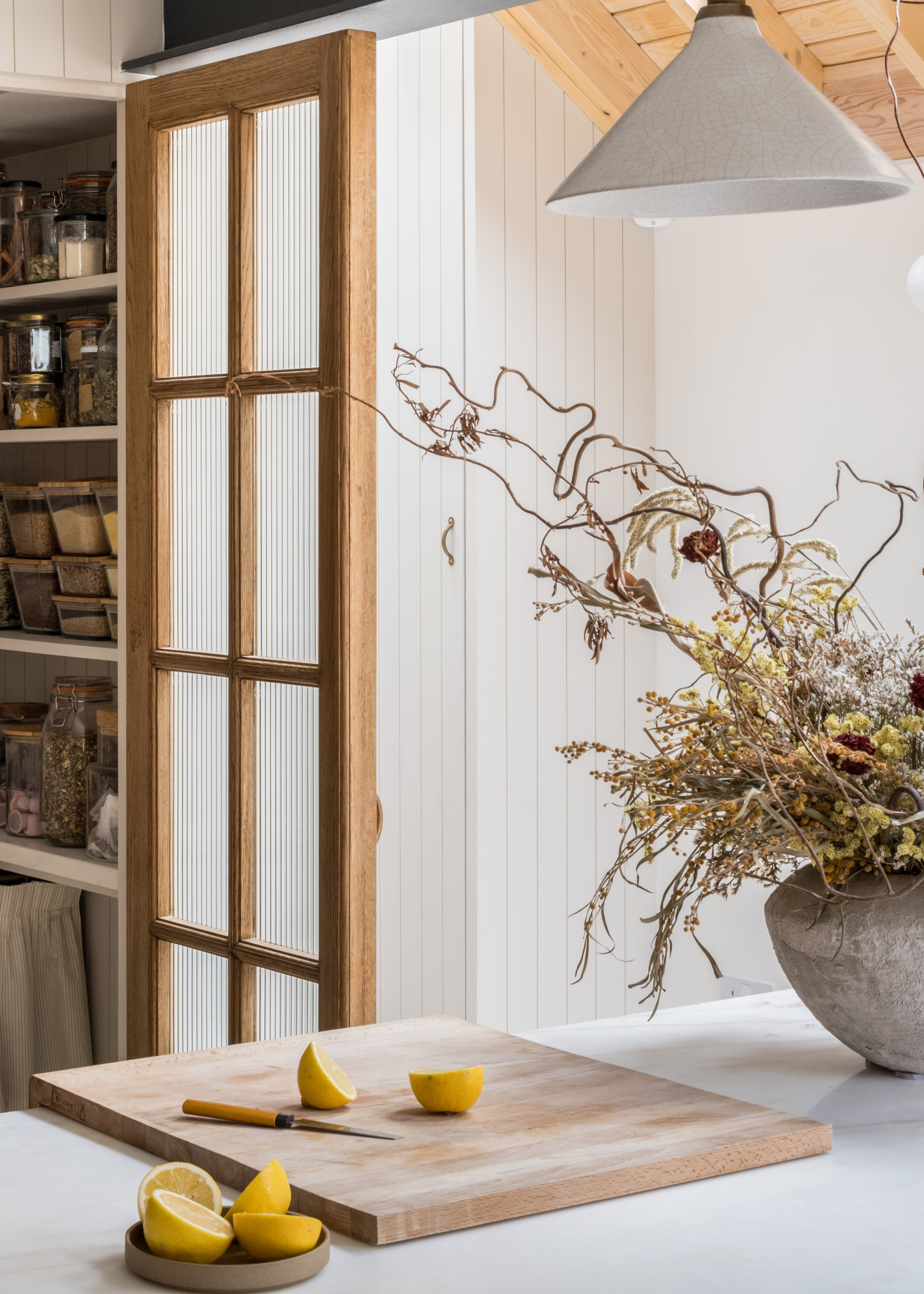 A kitchen with a wood pantry door a ceramic pendant light, a vase of dried stems and flowers by a chopping board with a bowl of sliced lemons on a stone island