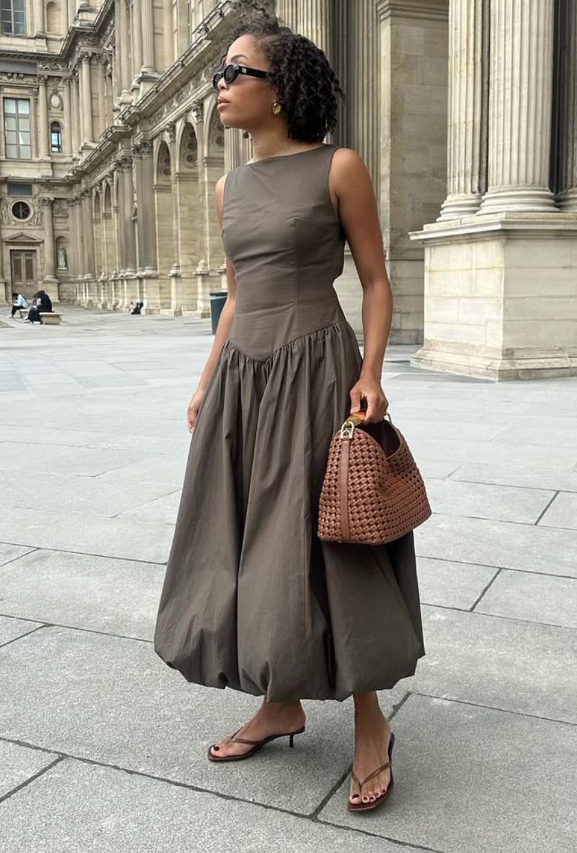 @slipintostyle trägt ein braunes Midikleid mit Ballonsaum, dunkelbraune Flip-Flops mit Absatz, eine Sonnenbrille und eine Clutch aus hellbraunem gewebtem Leder