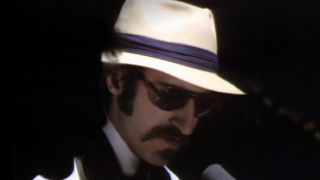 Leon Redbone performing with sunglasses and a hat on in darkness on SNL