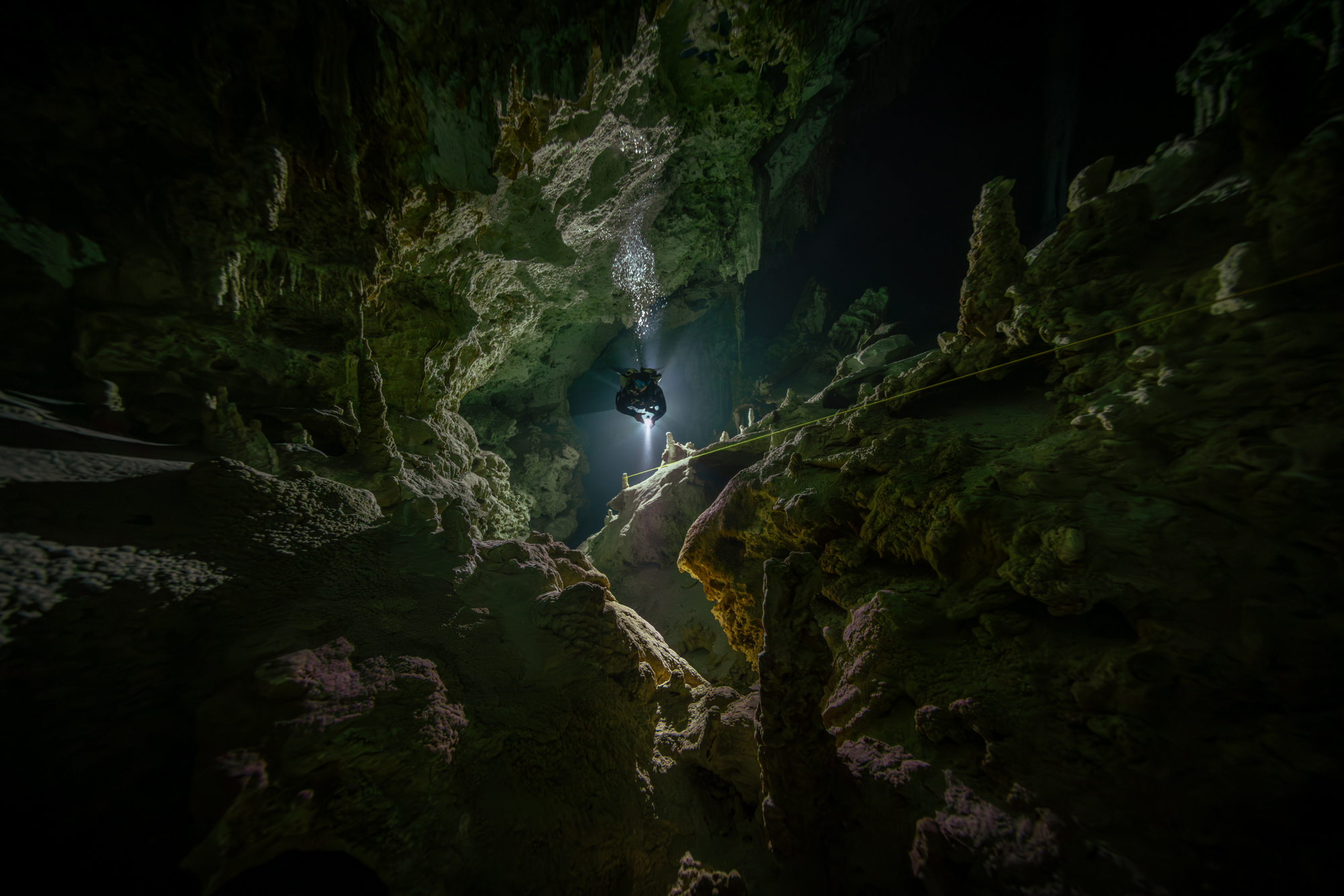 A lone caver hangs beneath a helmet light in a vast, moss-covered cave, illuminating jagged rock formations in a dark, mysterious cavern