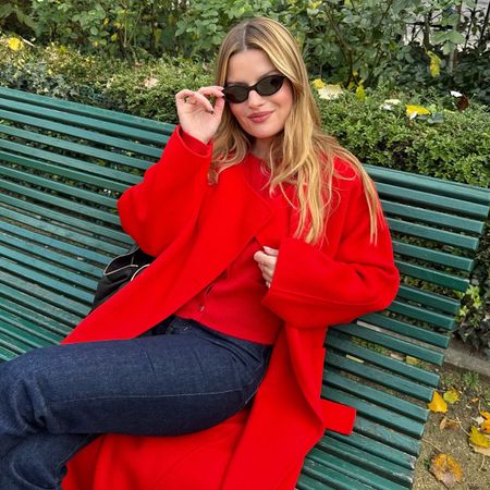 Sabina Socol wears a red cardigan under a red overcoat, dark-rinse denim, and a pair of black sunglasses. 
