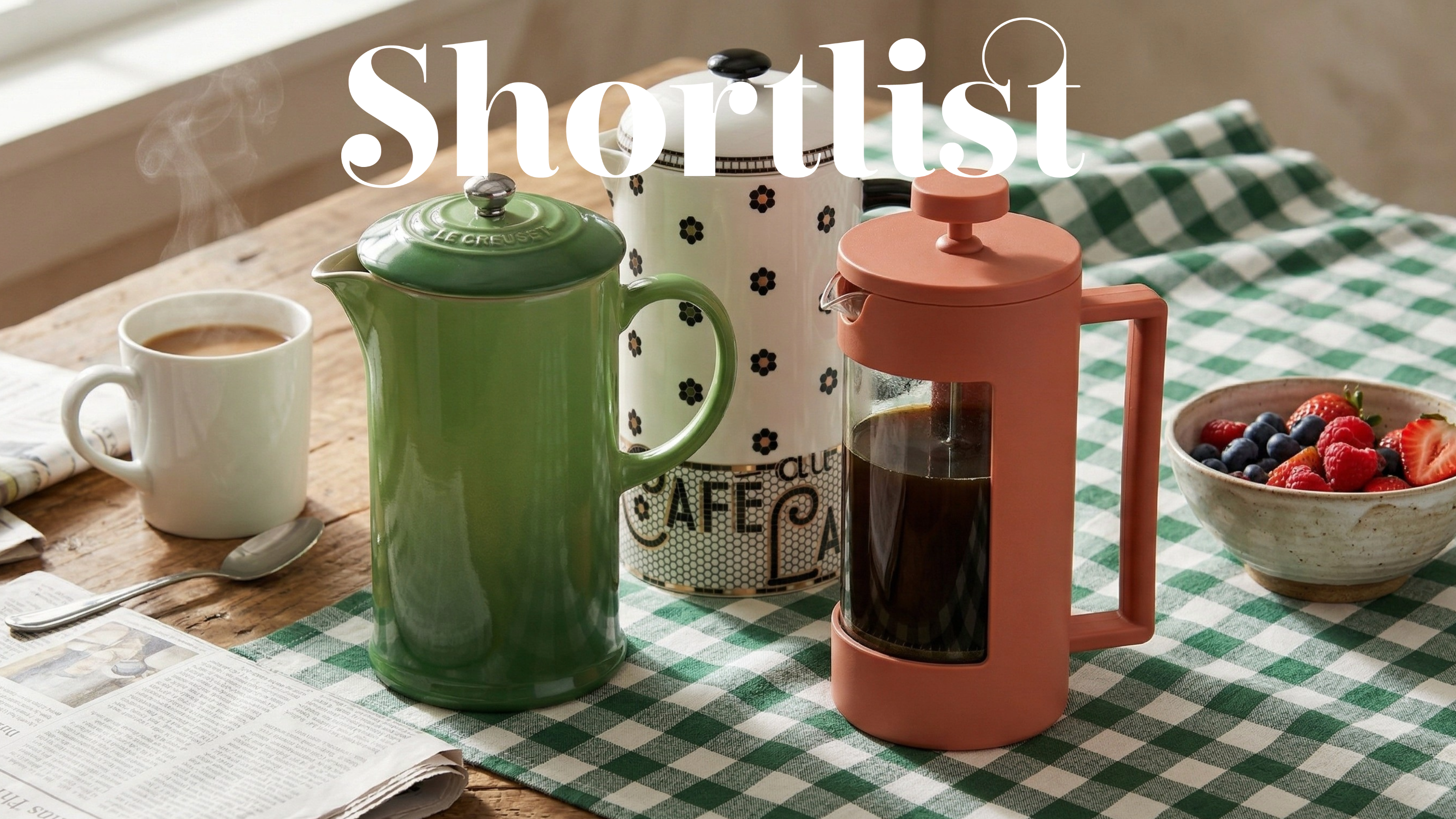 Three french presses on a gingham tablecloth with a bowl of fruit and a hot drink nearby 