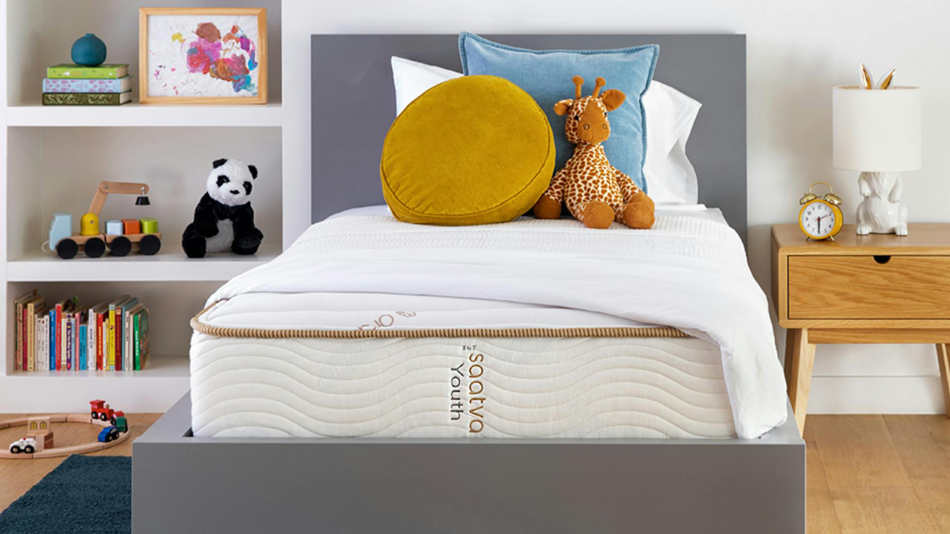 A front on shot of the Saatva Youth mattress on a bed frame in a kids room, some cushions and soft toys balanced at the head of the bed