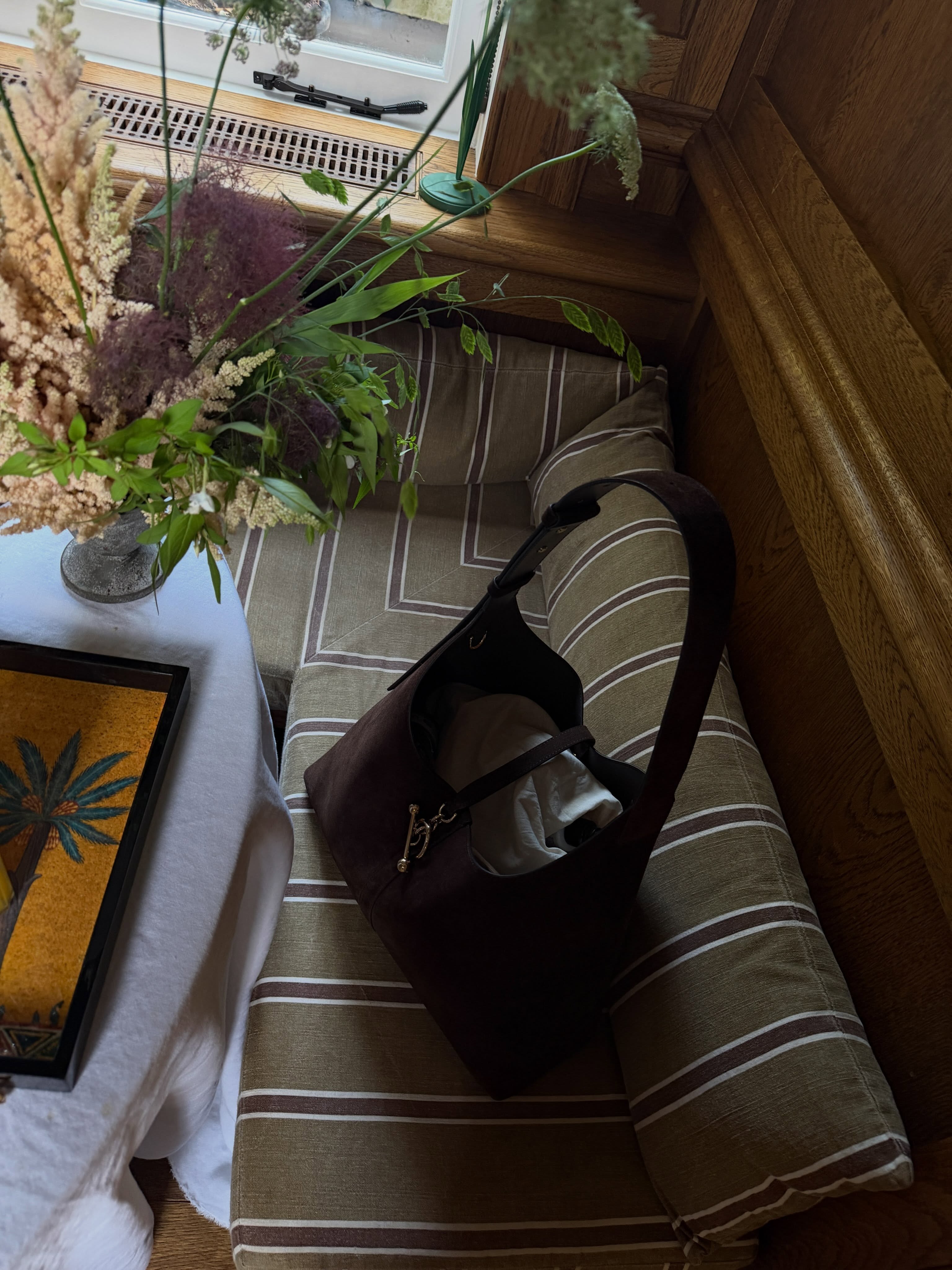 An image of a brown suede tote with a clasp closure on a striped green cushioned bench