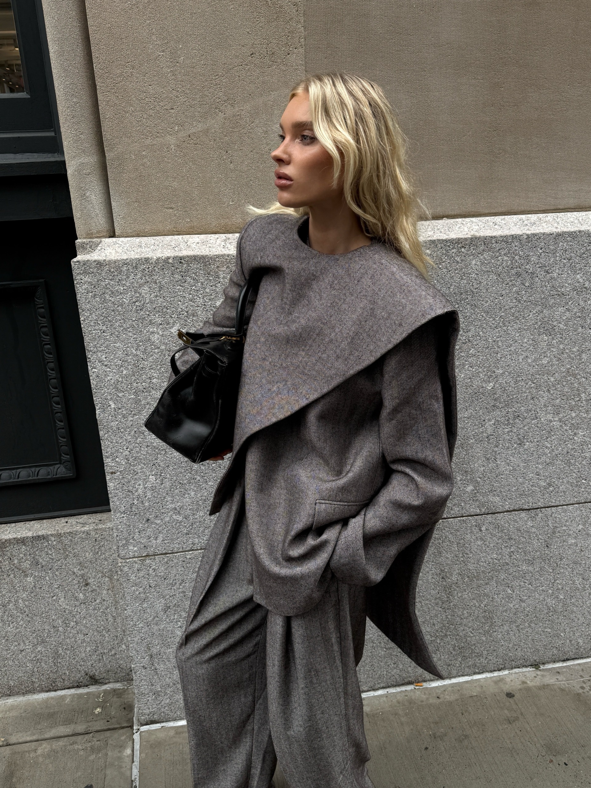 Elsa Hosk wears a grey scarf jacket with matching grey trousers in New York. She holds a black bag and wears black pointed-toe boots.
