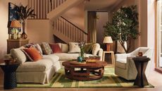A living room with a cream couch with colorful cushions, a wooden table with decor on it and a green rug beneath it, and a staircase in the background
