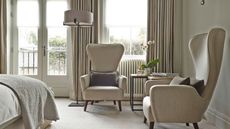 A carpeted cream bedroom with large upholstered chairs with brown cushions, large windows, curtains, a tall lamp, and a table with a flower and diffuser on it. In the left hand foreground, the corner of a bed is seen.