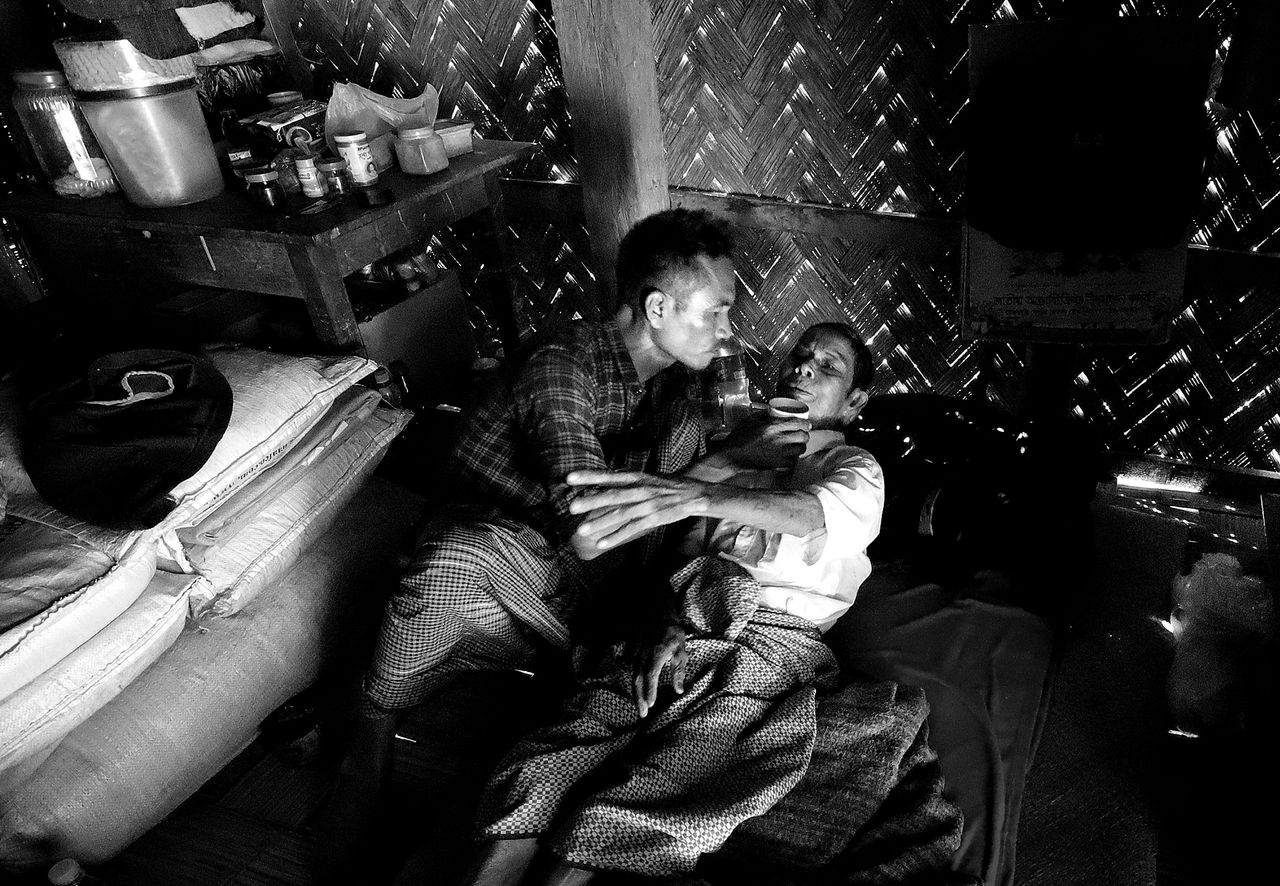 A dimly lit room with a woven wall, showcasing two individuals interacting closely on a bed, surrounded by household items
