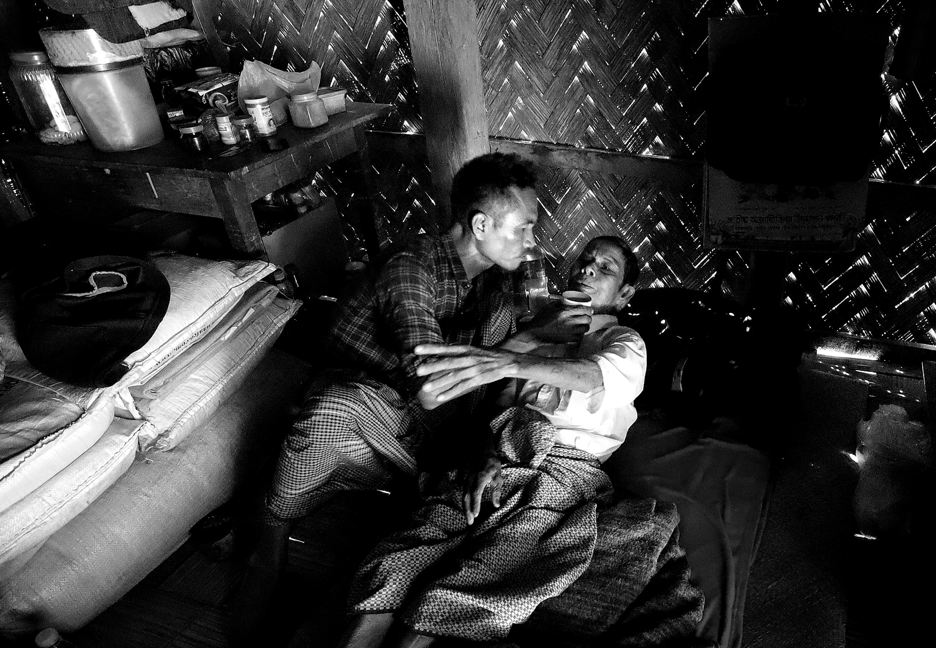 A dimly lit room with a woven wall, showcasing two individuals interacting closely on a bed, surrounded by household items
