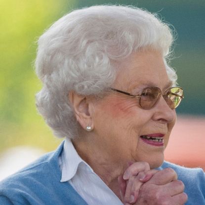 Queen Elizabeth wearing a blue sweater clasping her hands and smiling
