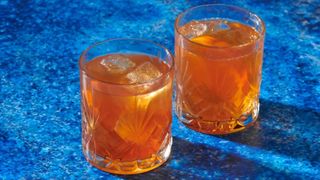 side shot of two beveled rocks glasses filled with orange liquid and ice against a shimmering blue counter