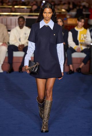 animal print boot trend is shown on the fall winter 2025 runway show of Tommy Hilfiger with a model wearing a blue button-down shirt under a navy dress with flat snakeskin knee-high boots and a black clutch bag