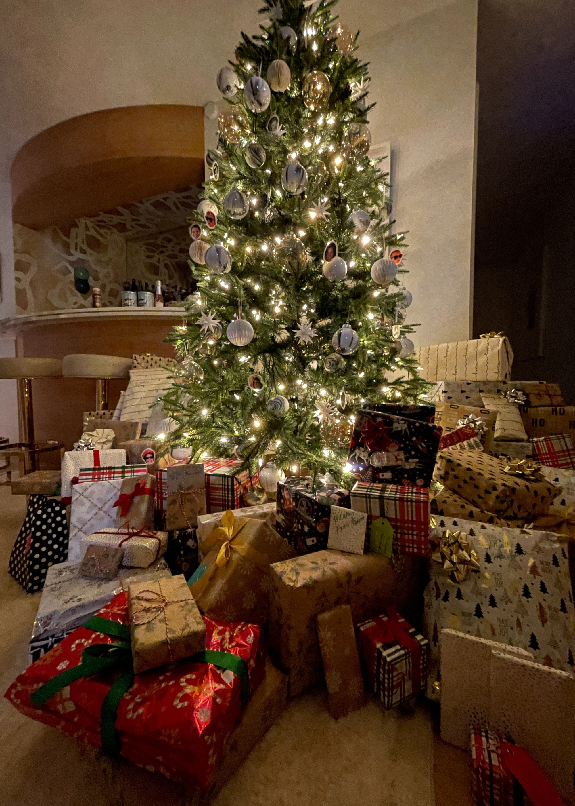 A lit and decorated Christmas tree with a huge mound of presents underneath and all around it next to a home bar