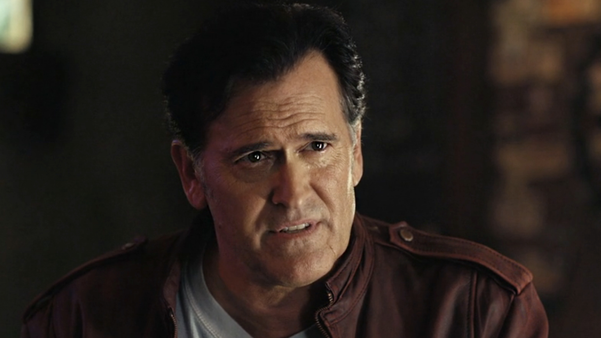 Ash (campbell) sits at the bar in a brown-red leather jacket and grey shirt in his return to his battle with the deadites in Ash vs Evil Dead.