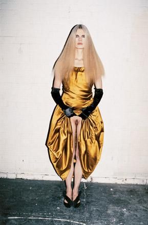 Lady with long blonde hair in centre parting wears a gold lame dress, black dress gloves and black shoes. The lady is lifting up her skirt to the waist revealing her lack of underwear.