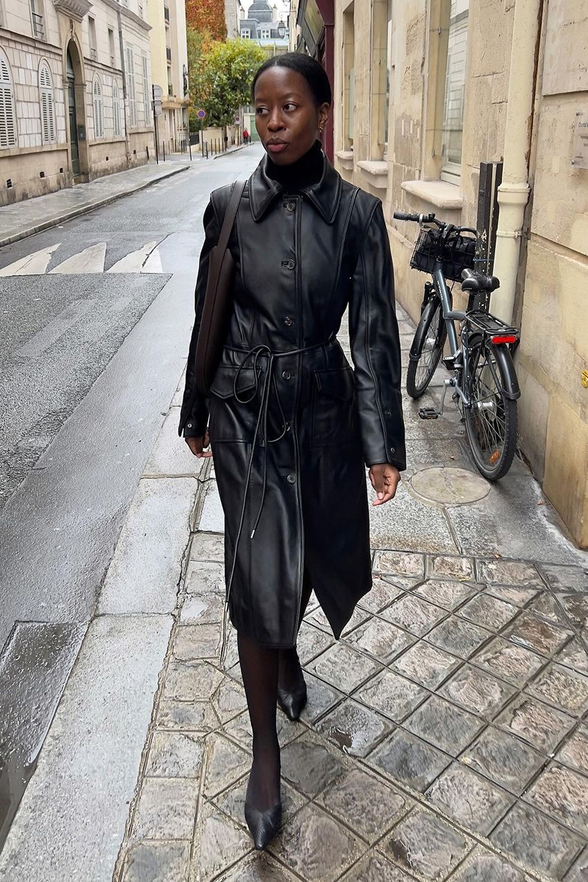 A woman walking down the sidewalk outside wearing a black duster jacket on top of a black turtleneck sweater, a pencil skirt, black tights, black pointed pumps, and a brown shoulder bag.