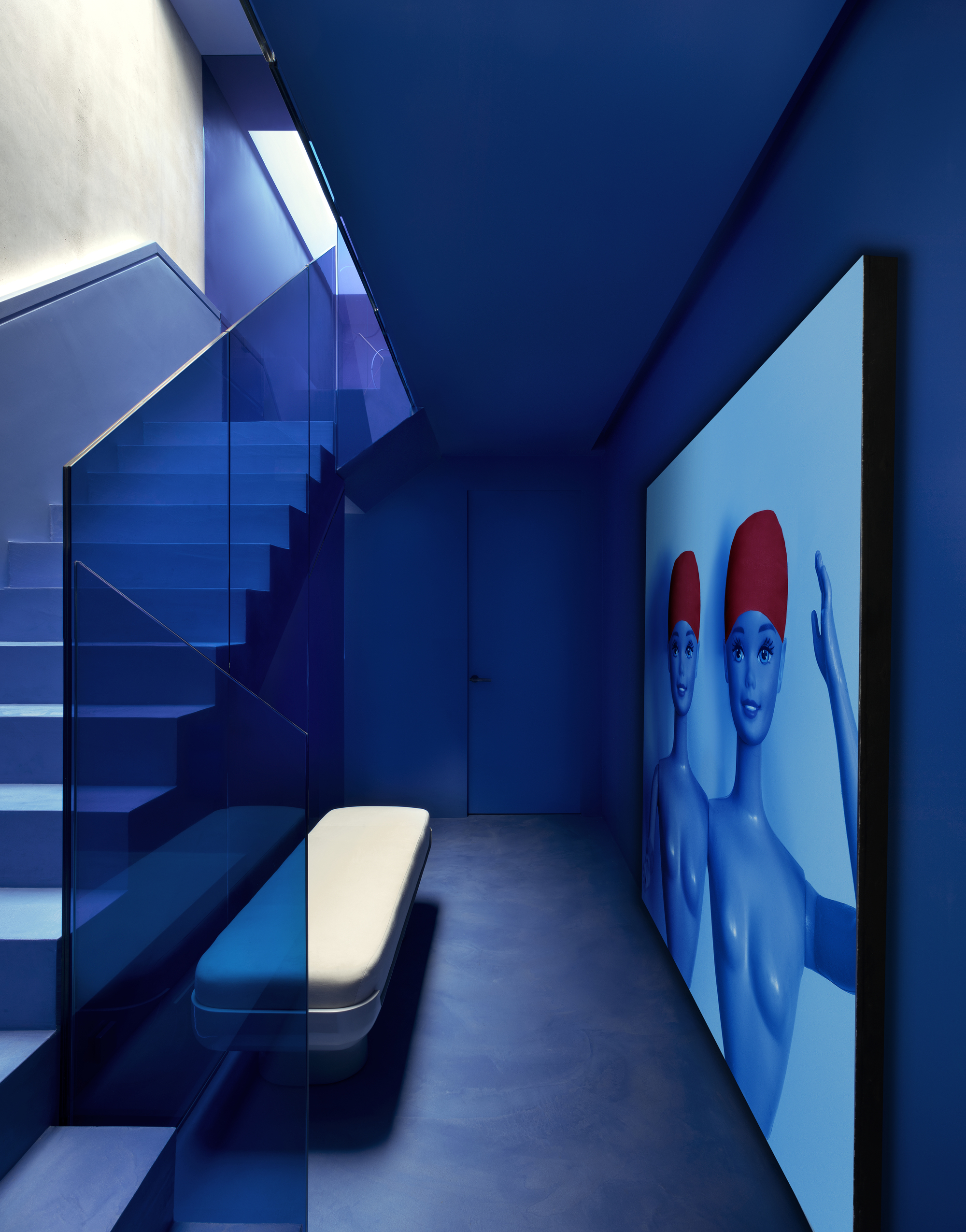 Image of a hallway and staircase drenched in a cobalt blue color. There is a white, modern bench seat at the base of the staircase and a large cobalt blue art piece of two Barbies with red swim caps.