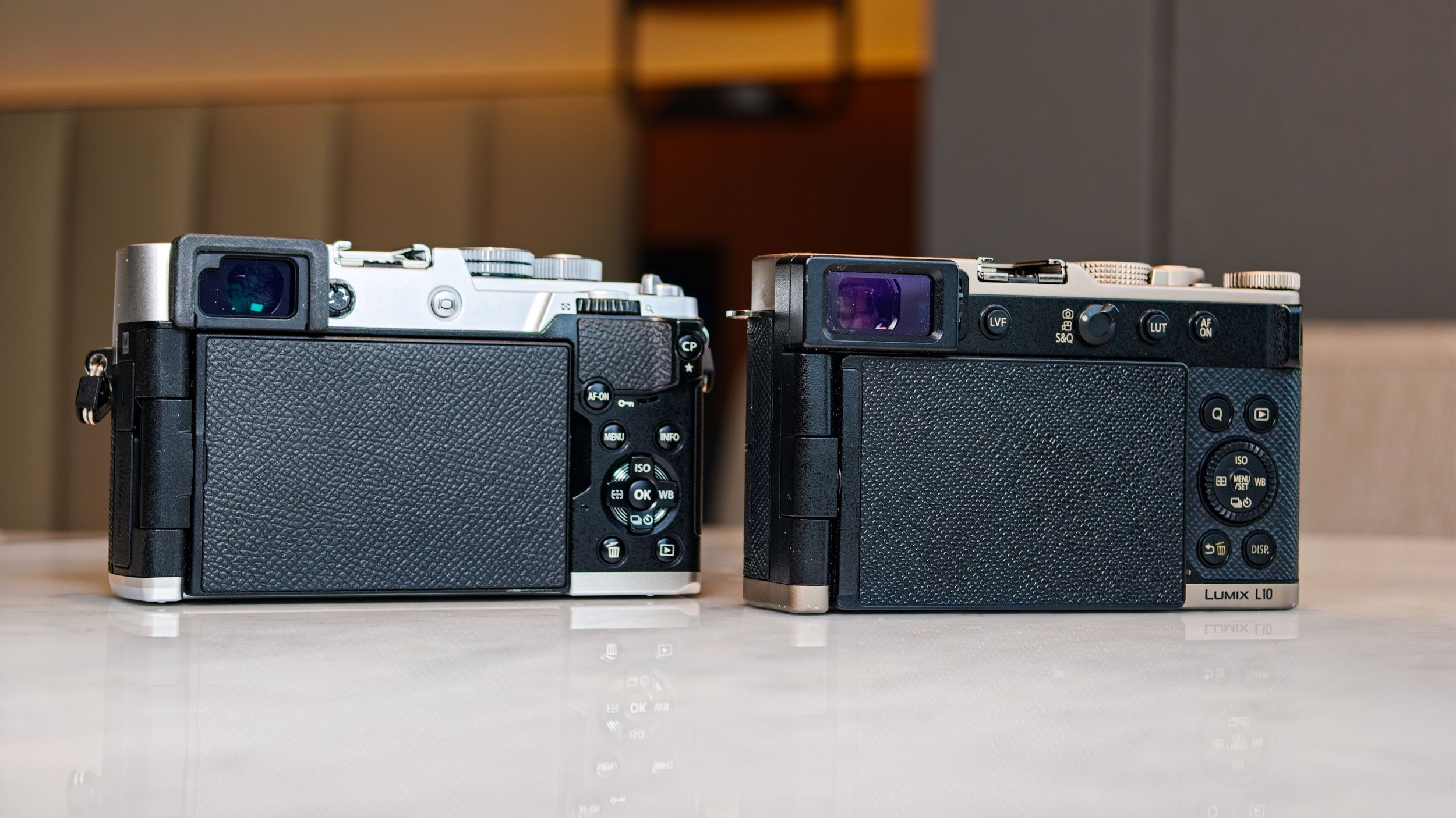 OM System PEN vs Panasonic Lumix L10: two micro four thirds cameras side by side on a white marble surface with lamp light in the background