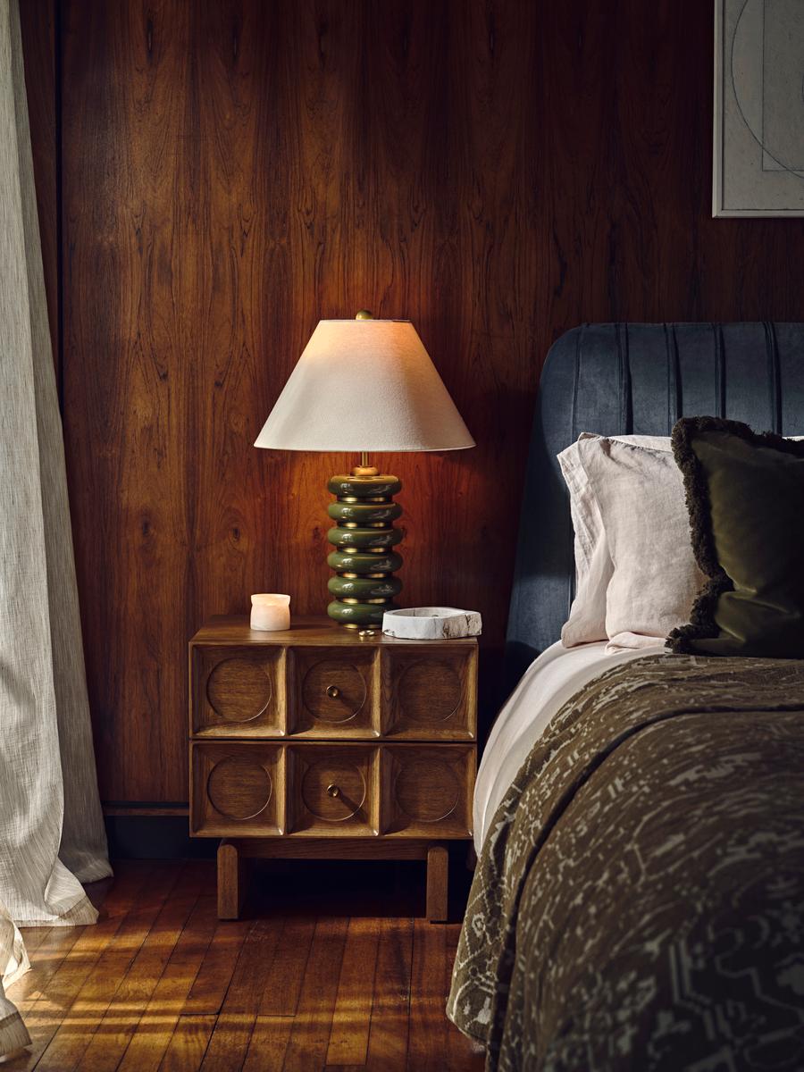 Bedroom with dark wooden walls, white curtains, wooden flooring, Marcel Bedside Table in Medium Oak and a green &amp;amp; white light on top, next to a bed with a blue headboard and dark green velvet bedding