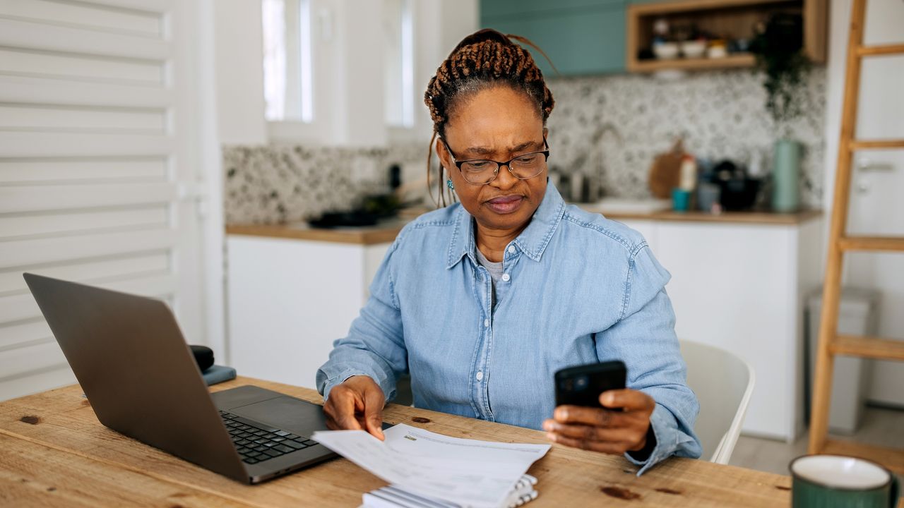 A retired woman uses a calculator while sitting with her laptop at her kitchen table.