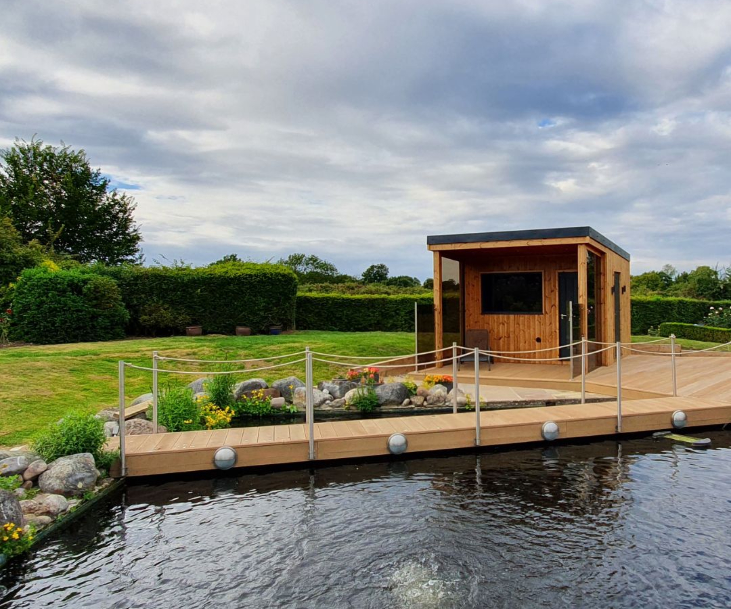 bespoke sauna built on wooden platform in large lawned garden with pond and wooden walkway across the water leading to sauna