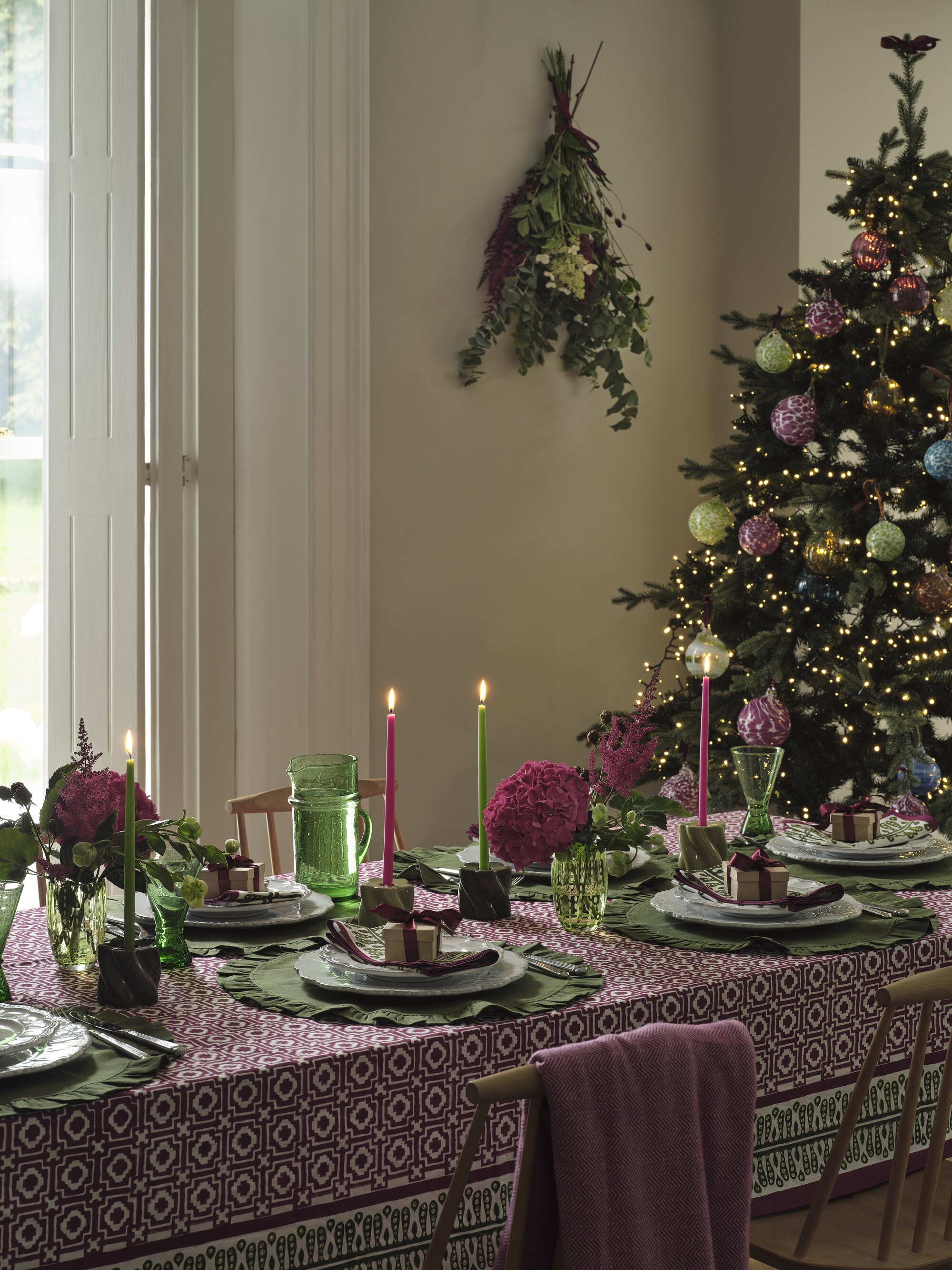 A dining table laid for Christmas with a purple patterned tablecloth, green fabric placemats, and a Christmas tree in the corner