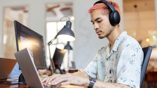 Male software developer using AI coding tools on a laptop computer while sitting at a desk with earphones on in an open-plan office environment.