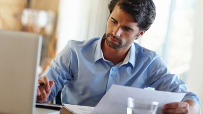 A man uses a calculator while holding paperwork and sitting in front of a laptop.