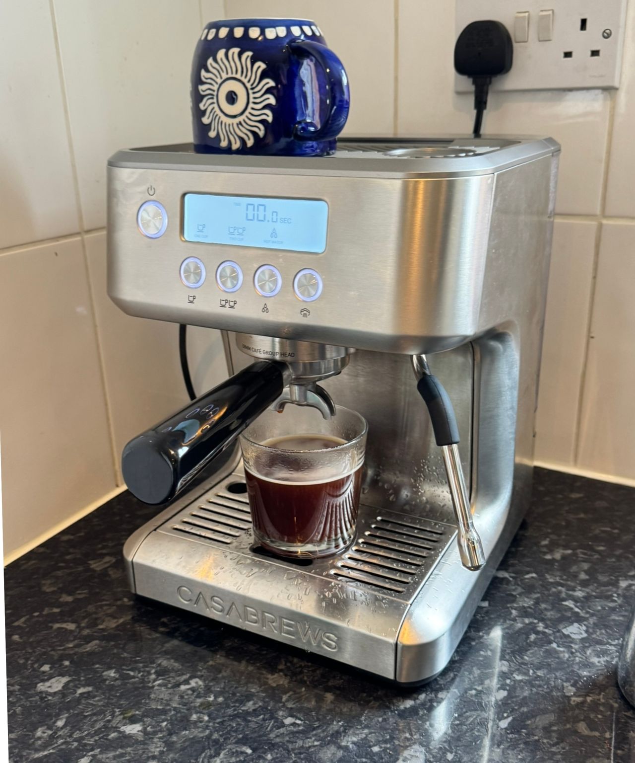 Casabrews ULTRA Espresso Machine on black countertop in front of white tiles, with blue mug positioned upside down on top of the coffee machine.