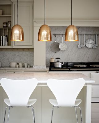 A grey kitchen with kitchen island, two white bar stools, three gold pendant lights over kitchen island and grey backsplash near hob with cooking equipment on rail