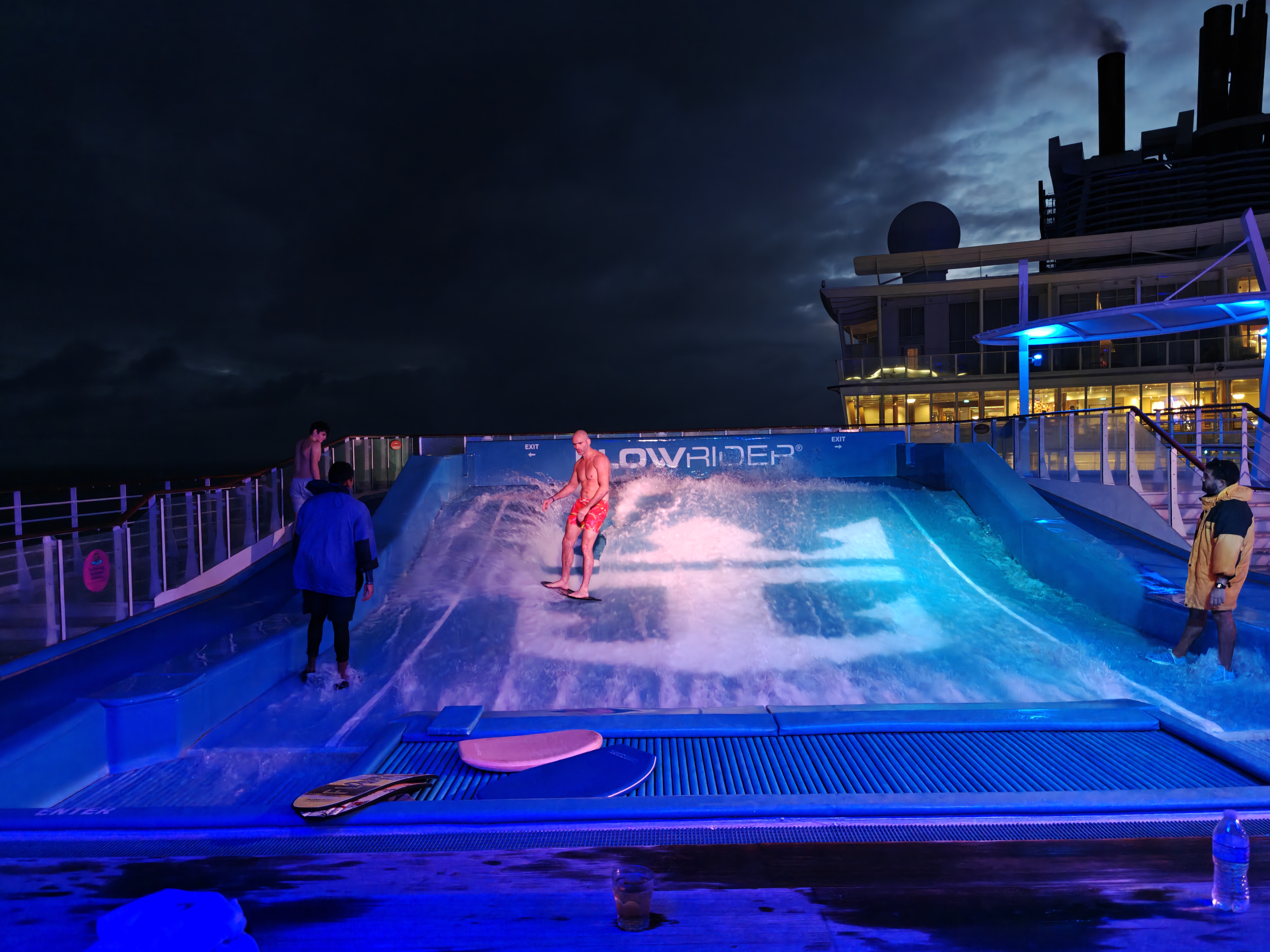 A man rides a surf board on the Flow Rider ride on a Royal Caribbean cruise at night with disco lighting