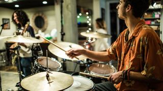 Man in orange shirt playing drums with a guitar player