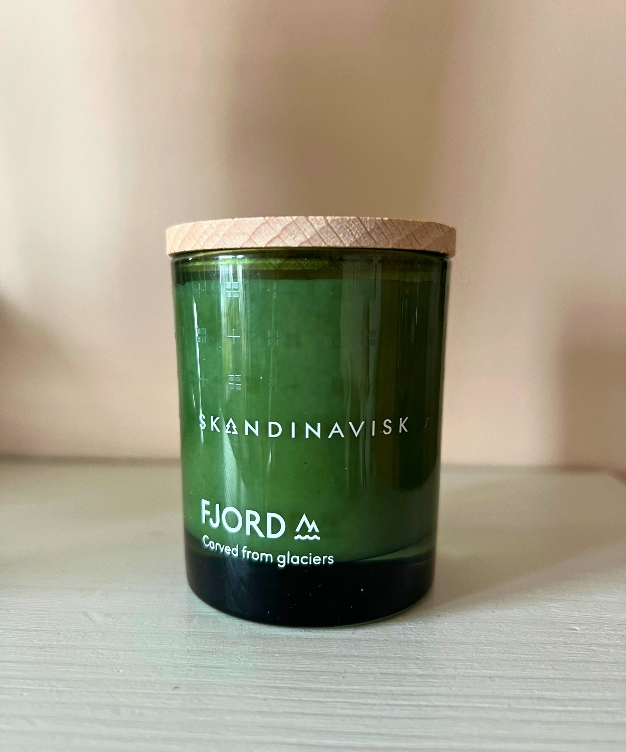 A green Skandinavisk candle with white lettering and a wooden lid on a white mantelpiece with a pink wall behind.