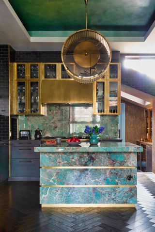 Marbled green kitchen with gold accents, kitchen island in the foreground and dark herringbone flooring.