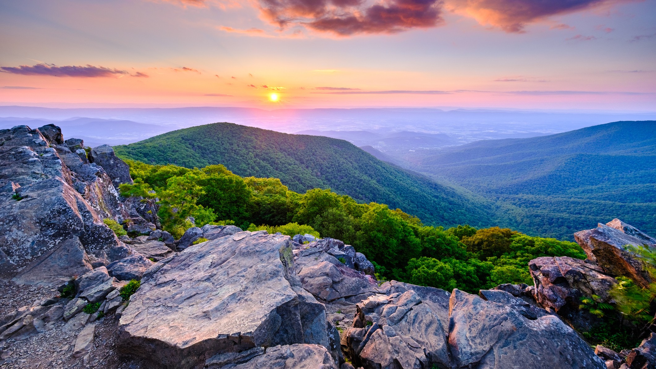 Sunset viewed from the summit of Hawksbill Mountain in Shenandoah National Park. We see mountains in the distance and rocks in the foreground.