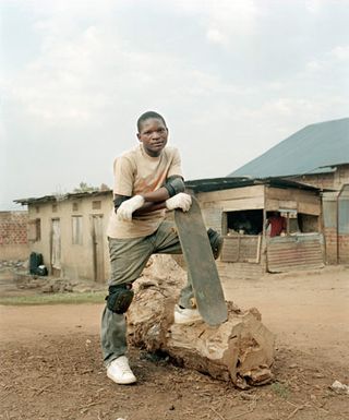An African man holding a skate board with one leg up on a tree stump.
