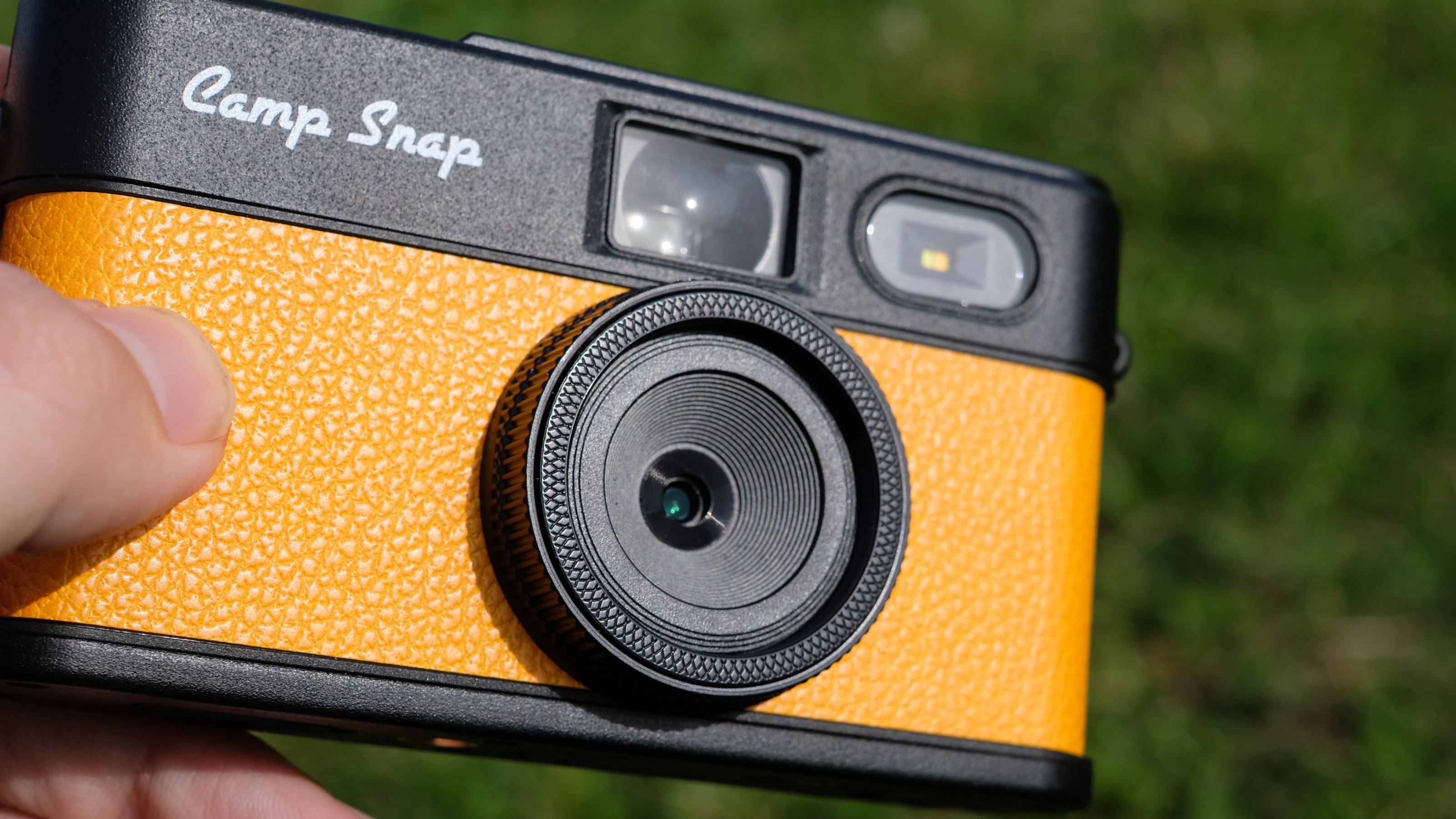 Photograph of man's hand holding Camp Snap 2 digital camera, in yellow colorway, with a closer look at the fixed lens with filter screw thread